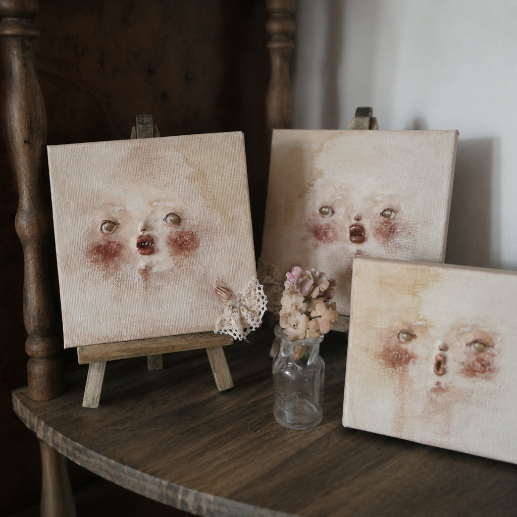 Three Lambwick clay-relief face artworks displayed on miniature easels on a wooden table.