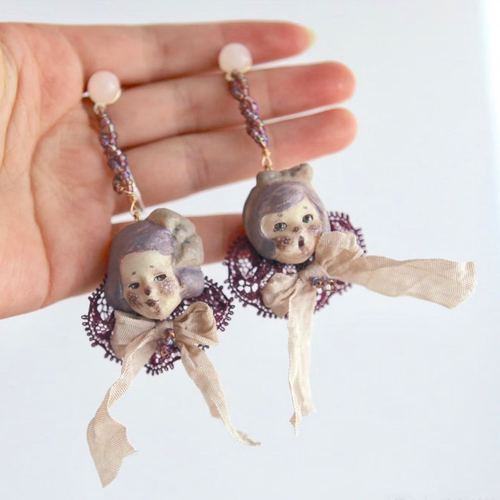 Mabel & Lucille doll-face earrings with lilac hair, purple lace and pale ribbons, held in a hand.