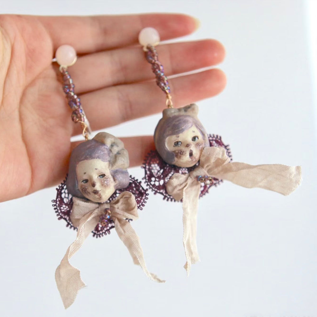 Mabel & Lucille doll-face earrings with lilac hair, purple lace and pale ribbons, held in a hand.