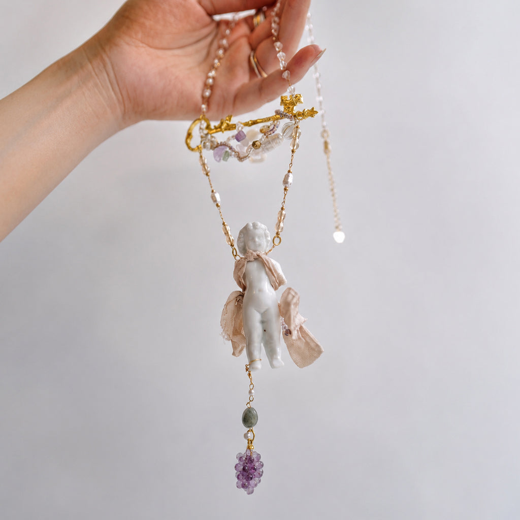 Charlotte ceramic-doll necklace held up to show the doll pendant, pink fabric and purple bead drop.