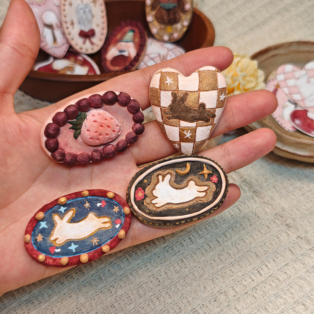 Winterwood strawberry, checked-heart and rabbit brooch designs displayed across a hand.