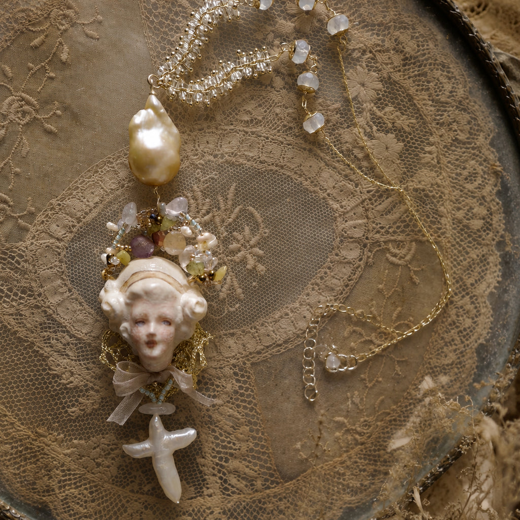 Close view of an Odette Cross doll pendant framed by beads and a large pearl-like accent.