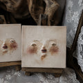 Two square Lambwick clay-relief faces with painted eyes and noses displayed on miniature easels.