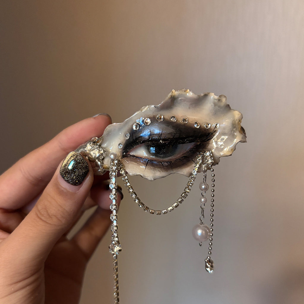 Lilith Veil Black & White Smoky oyster eye held in a hand, showing dark makeup and hanging chains.