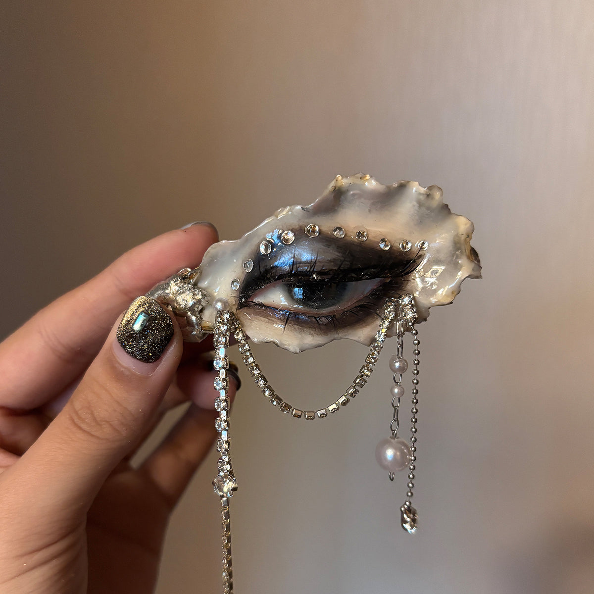 Lilith Veil Black & White Smoky oyster eye held in a hand, showing dark makeup and hanging chains.