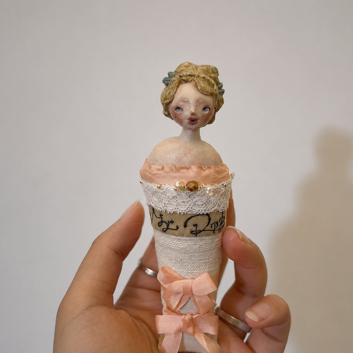 Lambwick Twin Series doll brooch with pale pink ribbons, held upright in a hand.