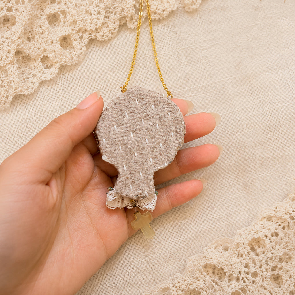 Stitched fabric back of a Lambwick doll-face pendant with a small cross drop, held in a hand.