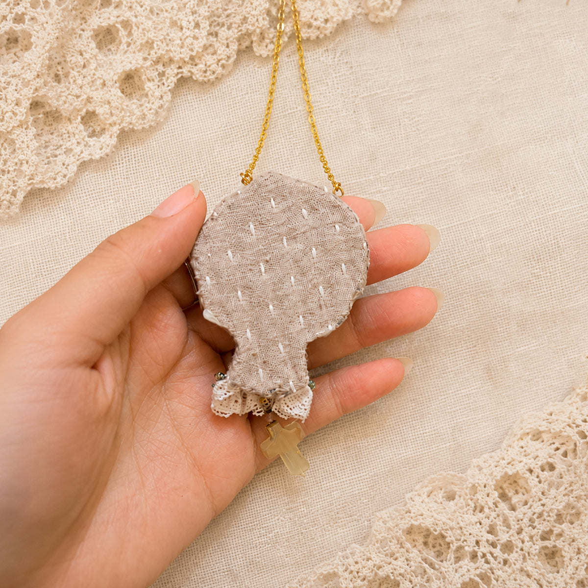 Stitched fabric back of a Lambwick doll-face pendant with a small cross drop, held in a hand.