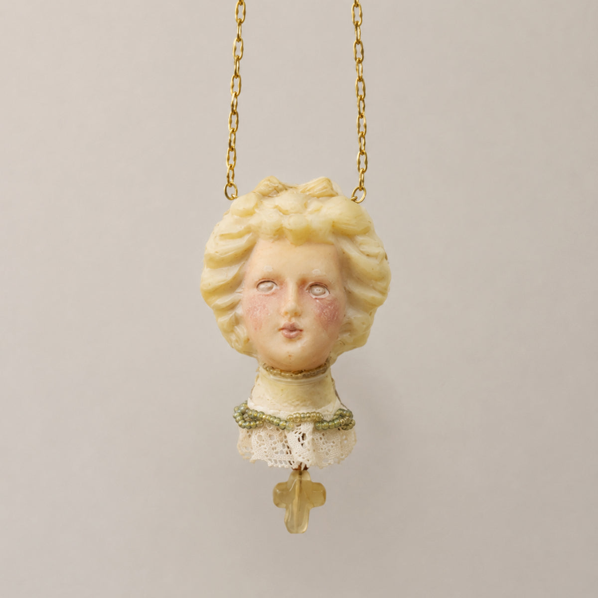 Blonde Lambwick doll-face pendant with a cream lace collar and a small cross drop.