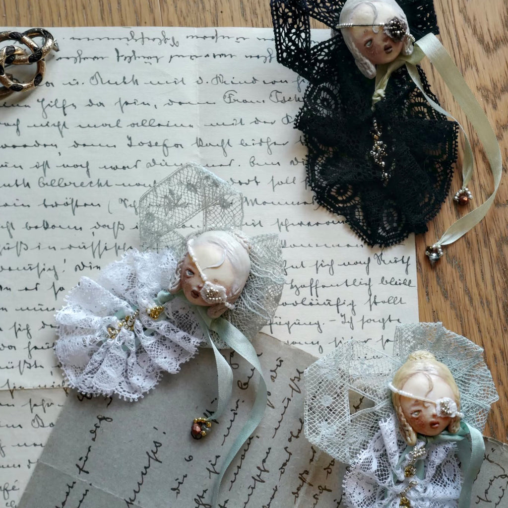 Black- and white-lace masked Lambwick doll brooches arranged together on handwritten paper.