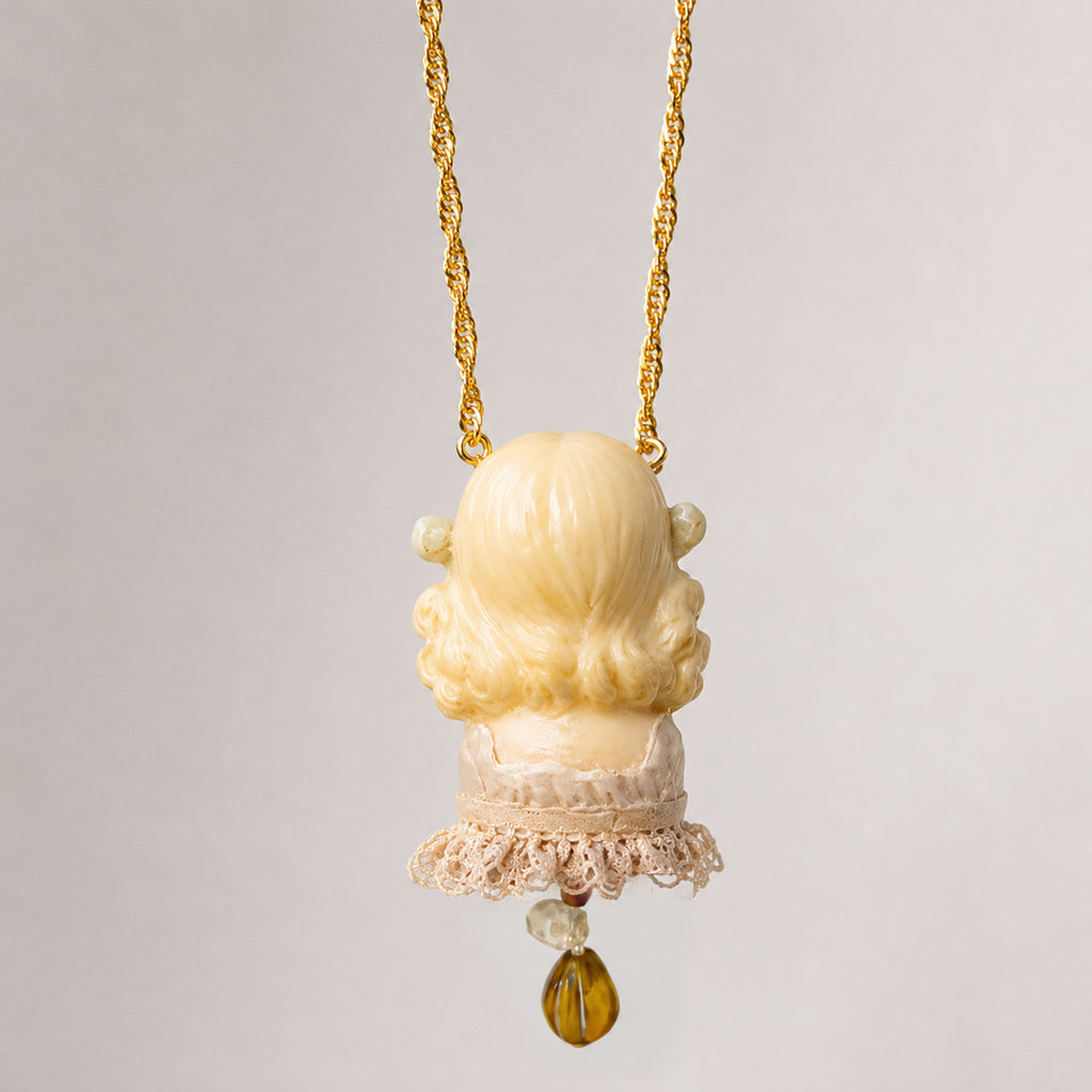 Rear view of a blonde Lambwick doll-head pendant with a lace collar and amber-colored drop.