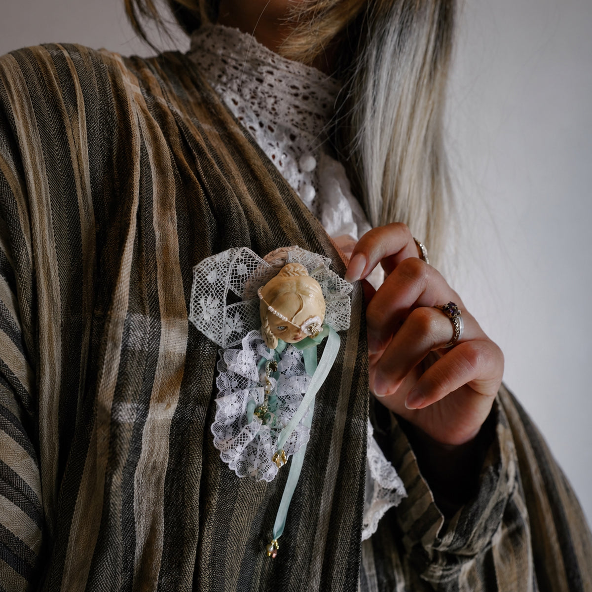 White-lace masked Lambwick doll brooch shown against striped fabric beside a hand.