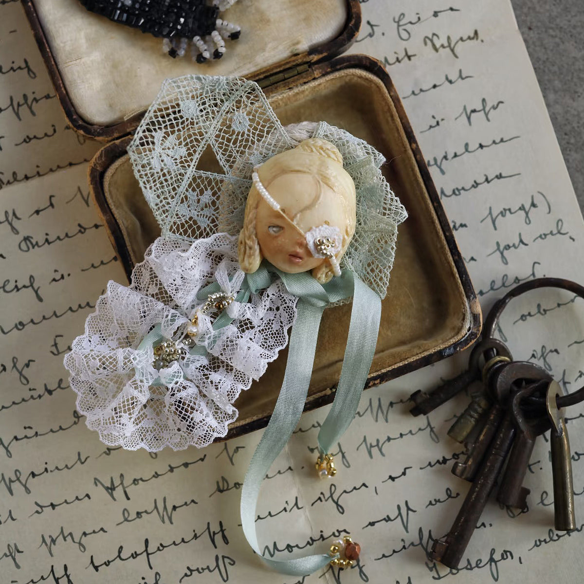 White-lace masked Lambwick doll brooch displayed inside an open box.