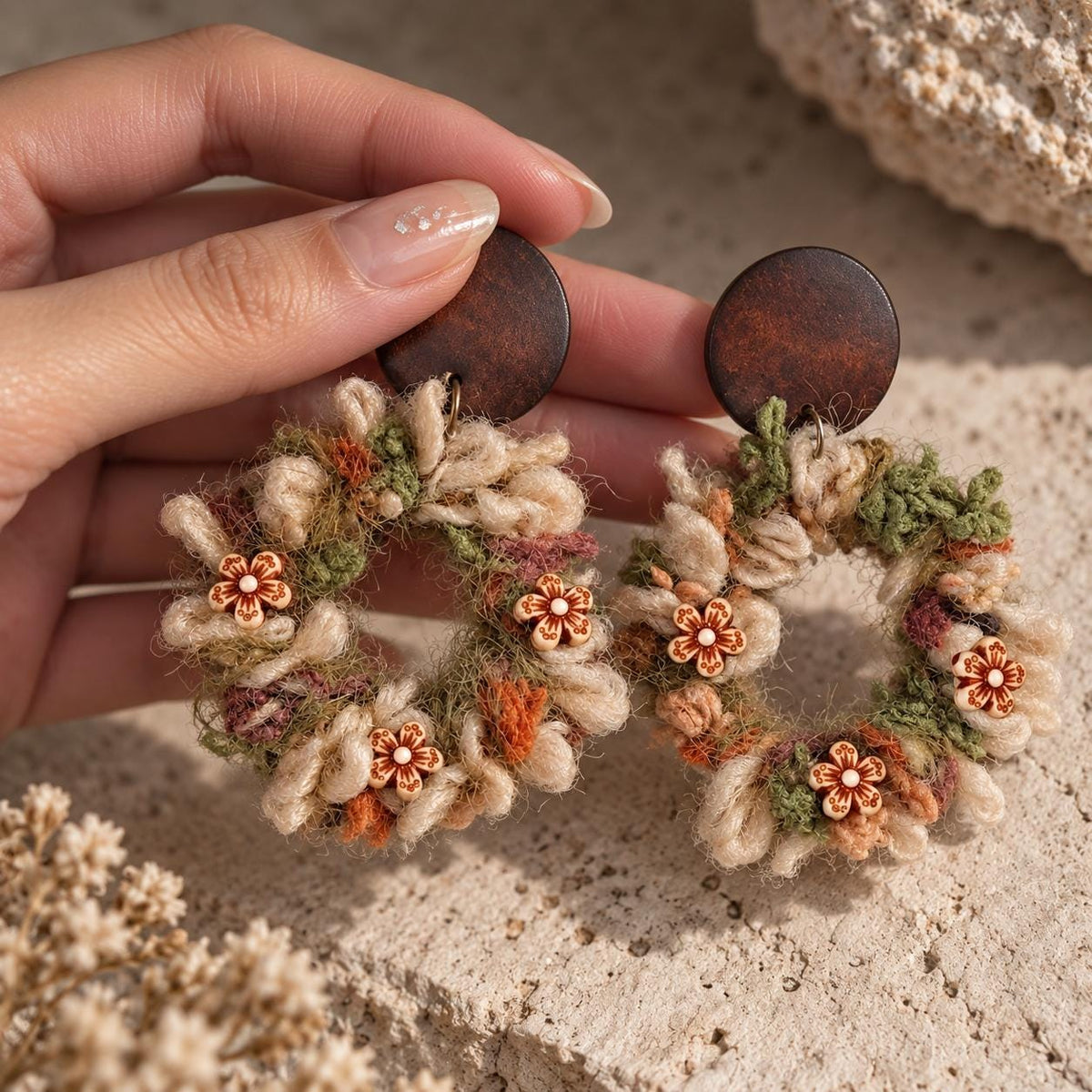 Pair of yarn floral-wreath earrings with brown studs held in a hand.