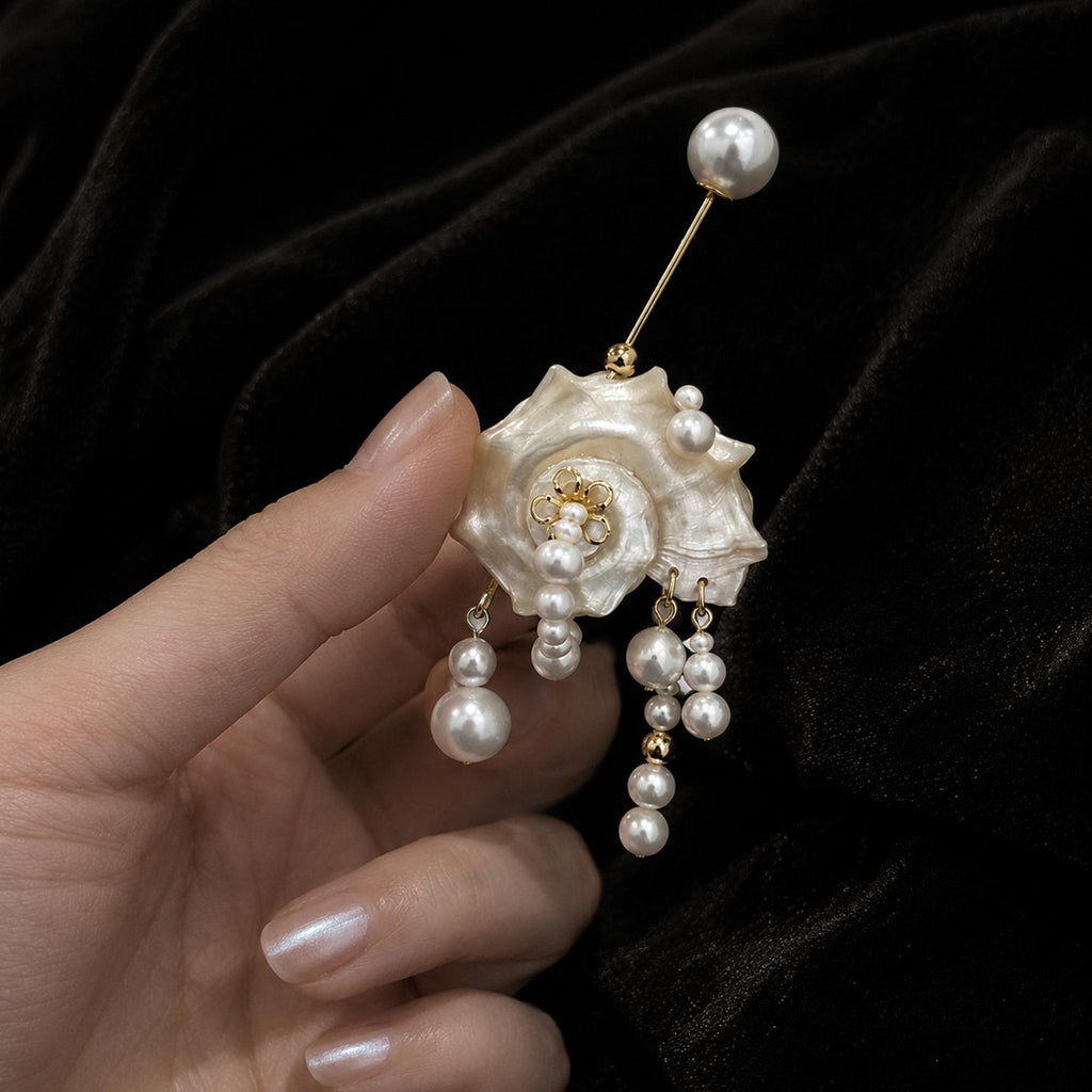 Conch-shell brooch with hanging beads and a pearl-topped pin, held in a hand.