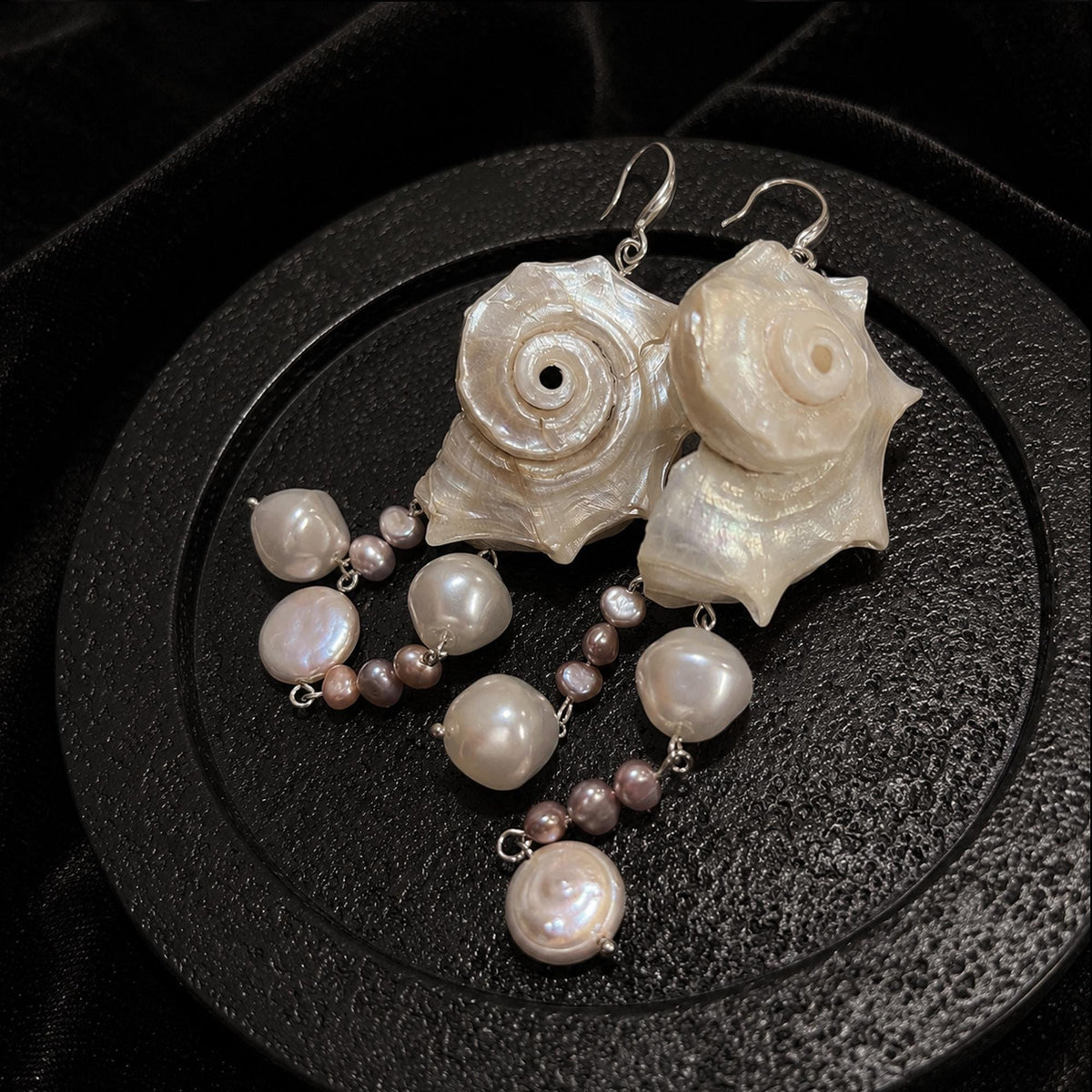 Spiral conch-shell earrings with pale and pink-toned bead drops on a round display stand.