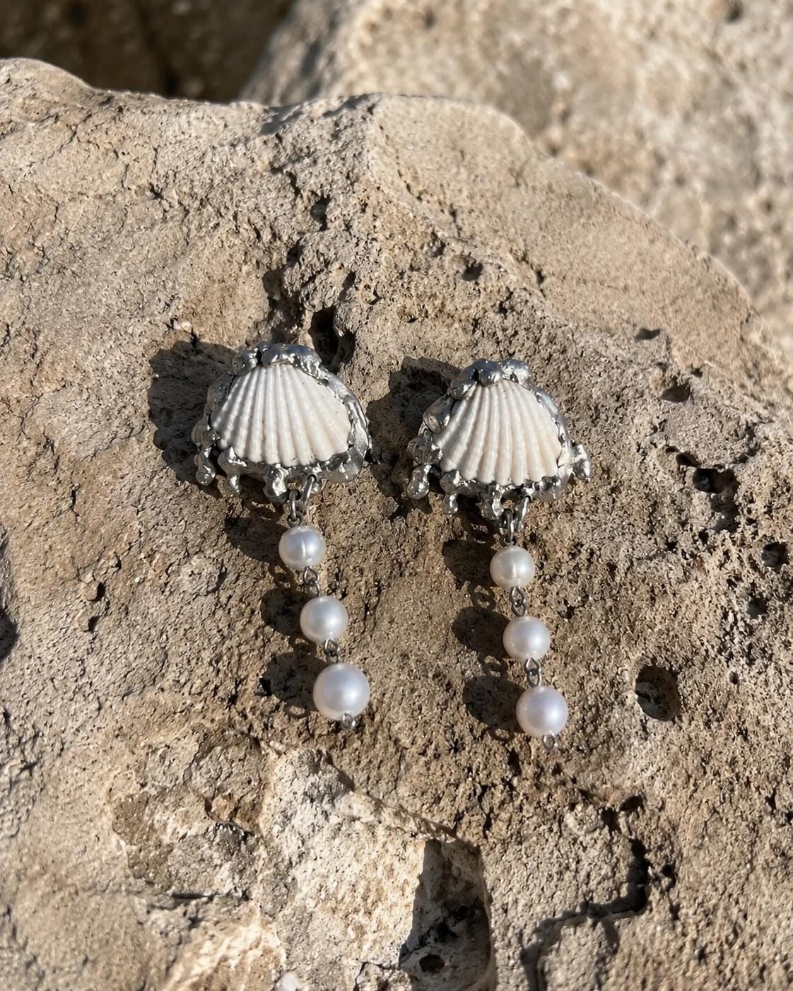 Two Shell Pearl Drop pieces with pale fan shells and hanging beads arranged on a beach rock.