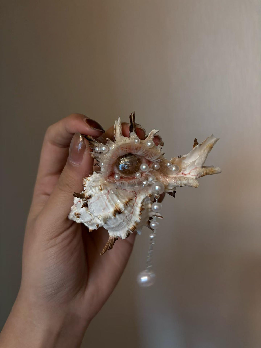 Pale spiked shell-eye sculpture with a hanging pearl-like drop, held in a hand.