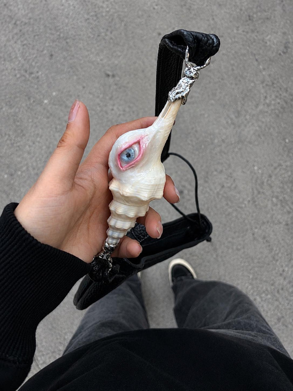 Close-up of the Long Conch Eye Shoulder Bag strap and pale shell-eye ornament, held in a hand.