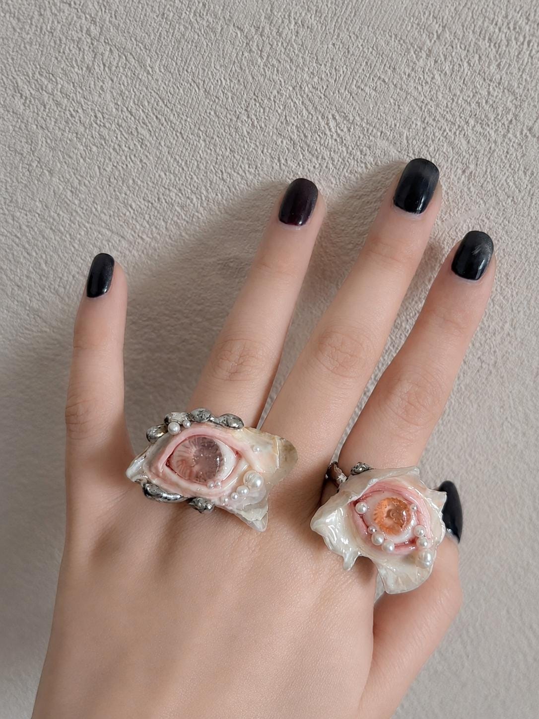 Two pale shell-eye statement rings with pink eyelids shown on a hand against a light wall.