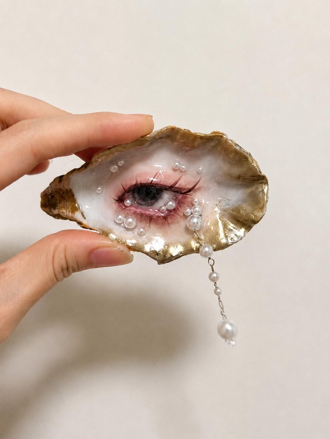 Gilded Oyster Eye held against a light wall, showing a hanging pearl-like drop.