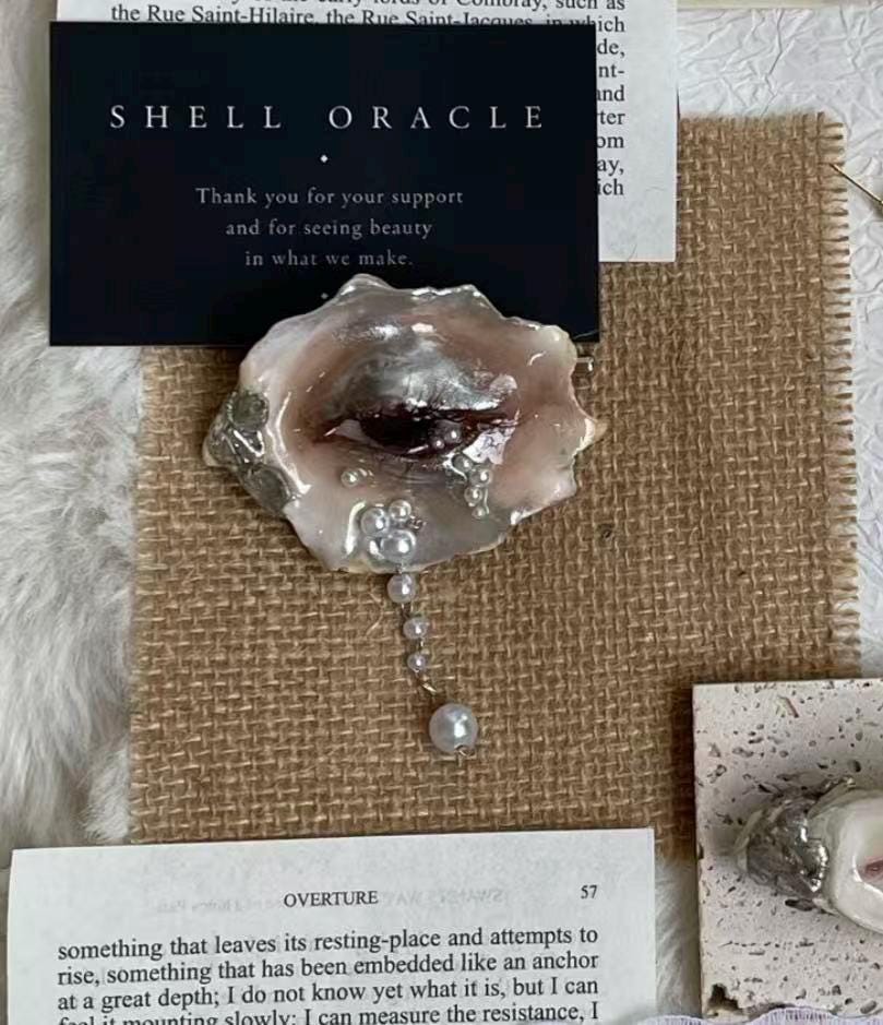 Brown Smoky Oyster Eye brooch displayed on printed paper beside a Shell Oracle card.