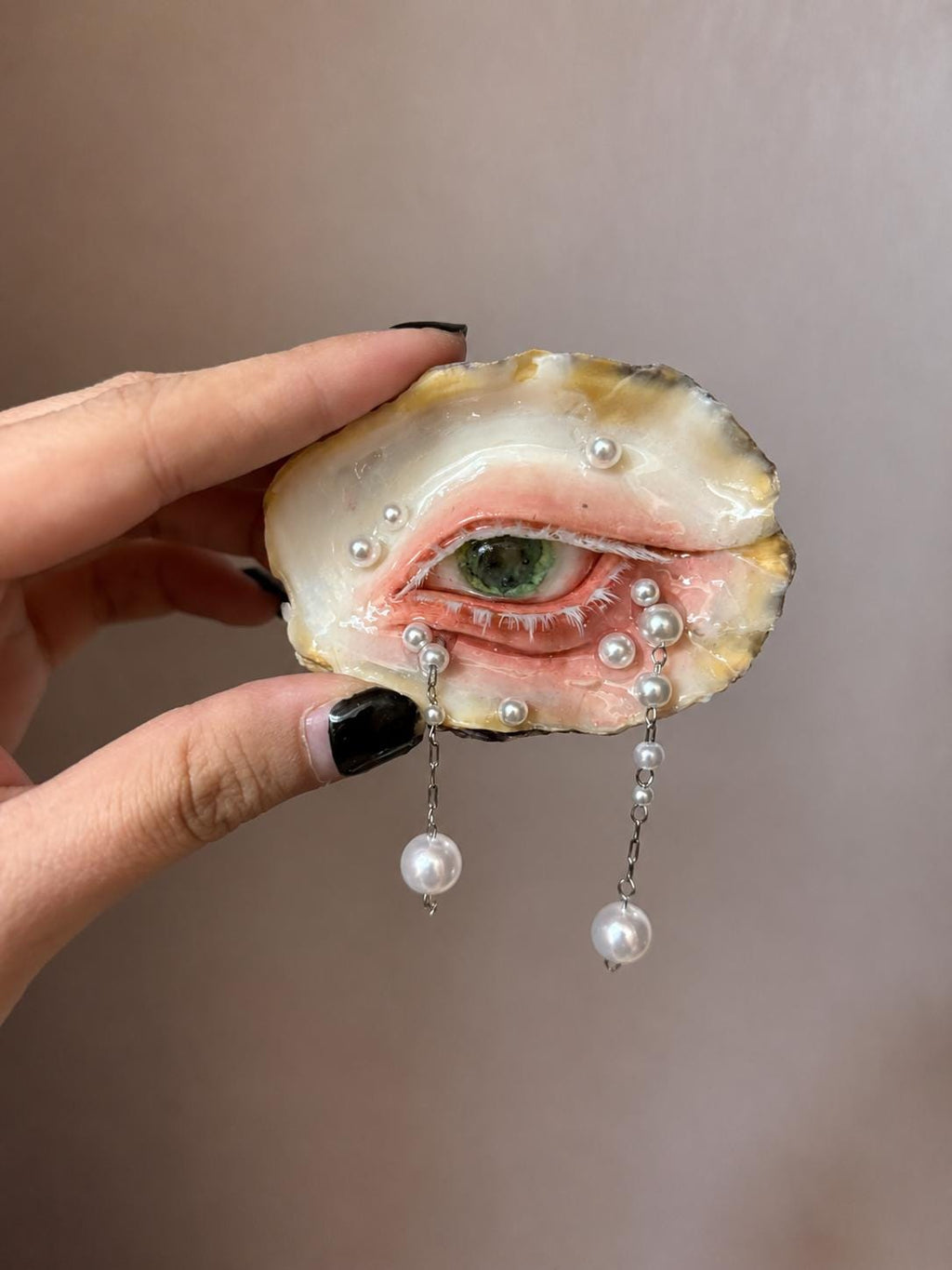 Larger Green-and-Gold Oyster Eye held in a hand, showing a green iris and hanging pearl-like drops.