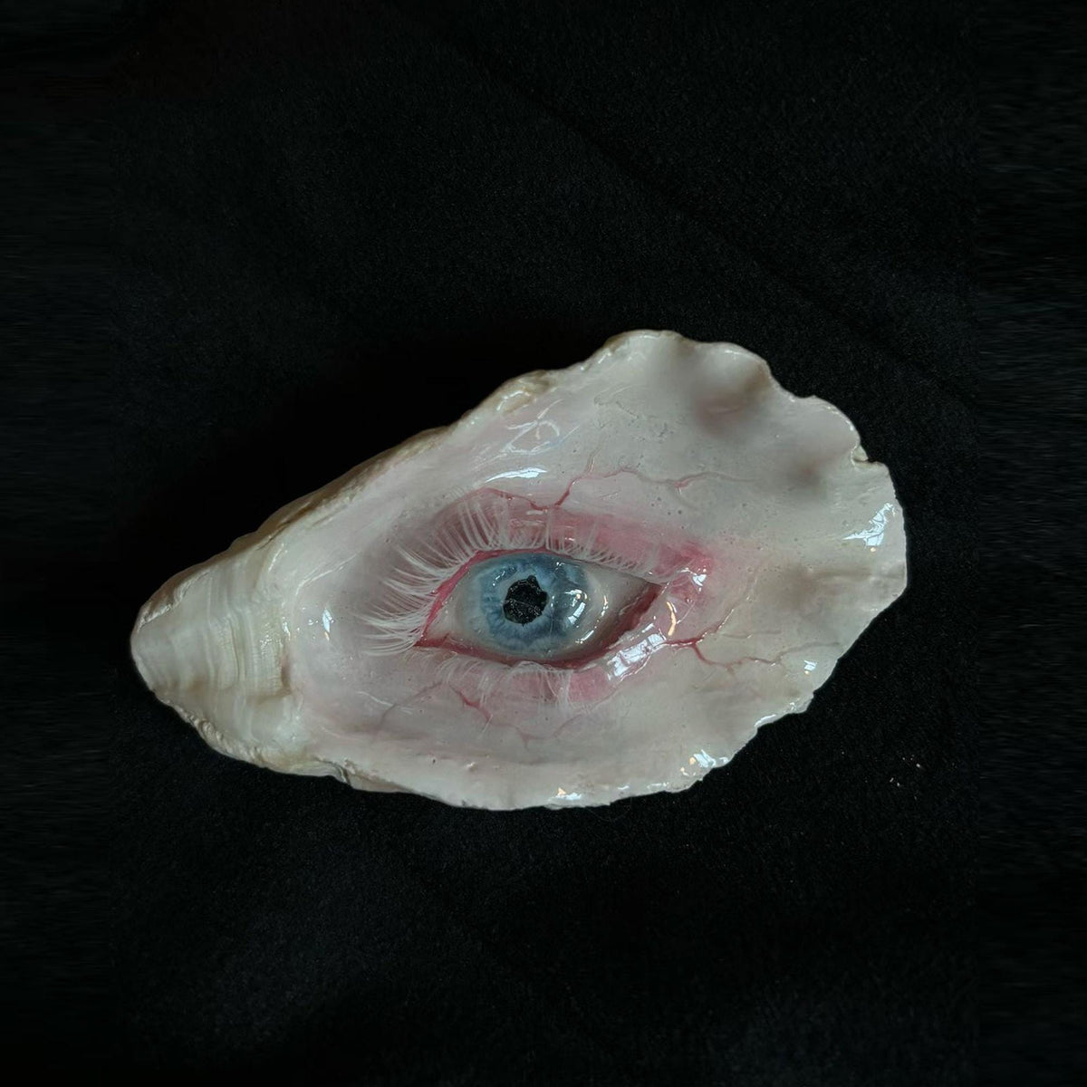 Small shell-eye artwork with a blue iris and painted red veins resting on dark fabric.