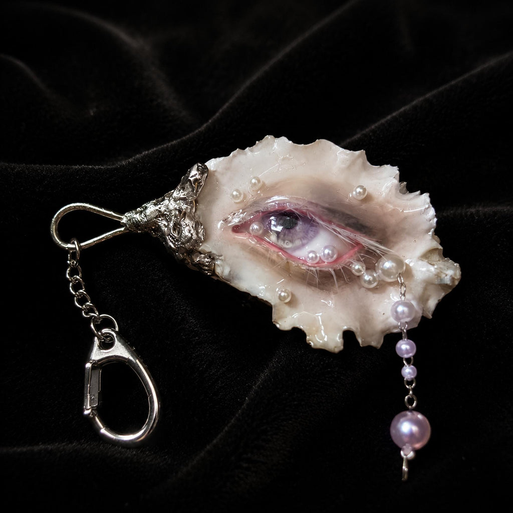 Nyra Ash Grey-Lash oyster eye keychain on black velvet with a hanging pearl-like drop.