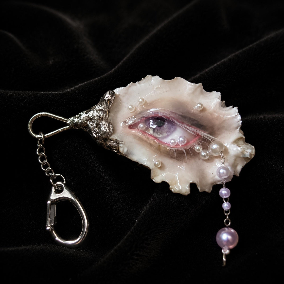 Nyra Ash Grey-Lash oyster eye keychain on black velvet with a hanging pearl-like drop.