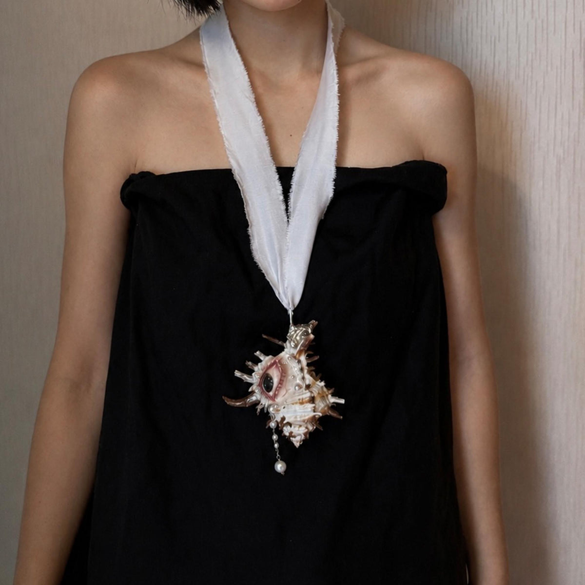 Abyss Guardians shell-eye necklace hanging from long ivory ribbon over a strapless black dress.