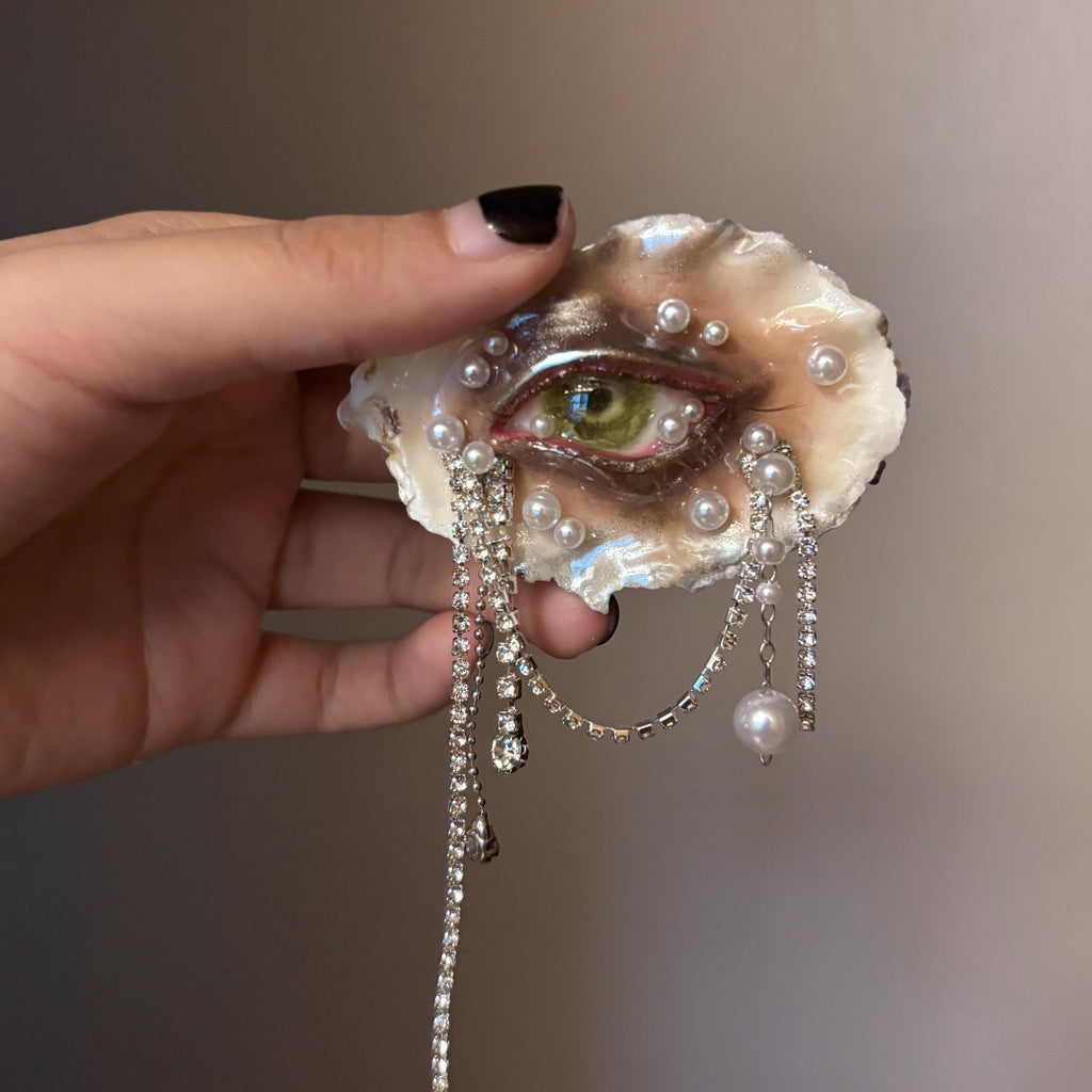 Lilith Veil Emerald Siren oyster eye held between fingers, showing green iris and hanging bead strands.